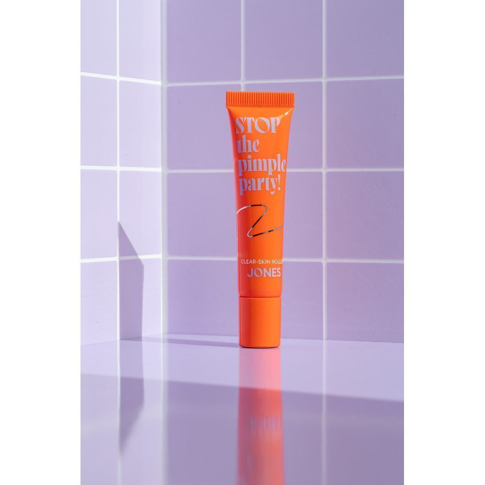 Oranje tube met roll-on applicator, met de tekst "STOP the pimple party! JONES" op een paarse tegelachtergrond.