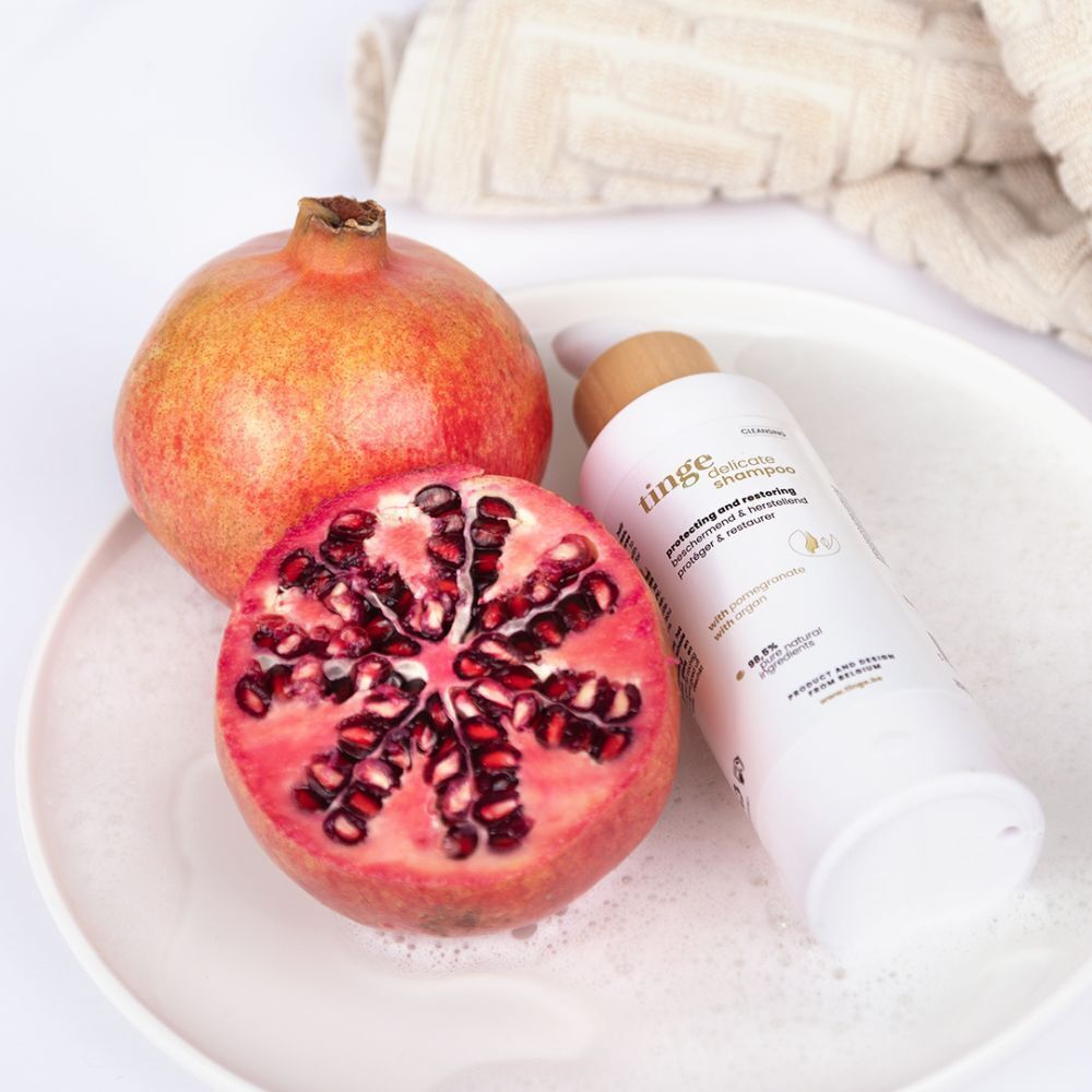 Een fles Tinge Delicate Shampoo naast granaatappels op een bord met schuim.