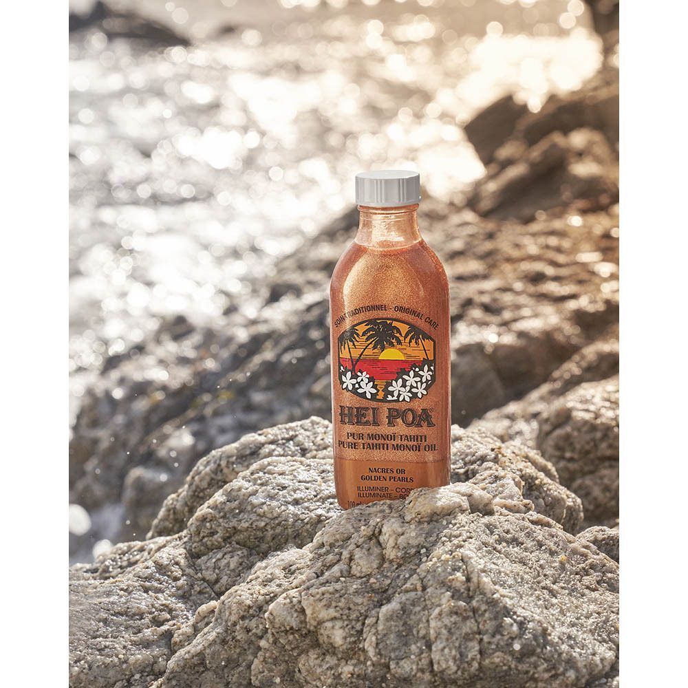 Fles Hei Poa Pur Monoï Tahiti met gouden parels op rotsen aan zee.