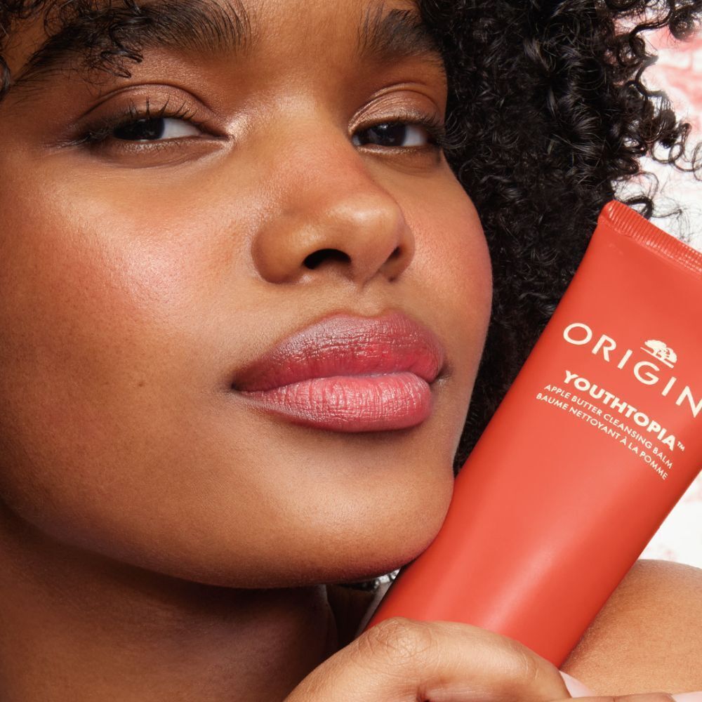 Gezicht dat een tube Origins Youthtopia Apple Butter Cleansing Balm vasthoudt.
