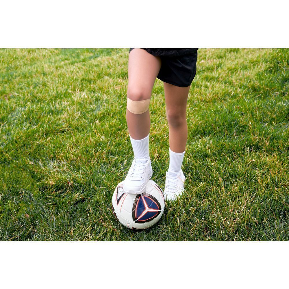 Enfant avec pansement Nexcare Ultra Stretch MAXI sur le genou, debout sur un ballon de football.