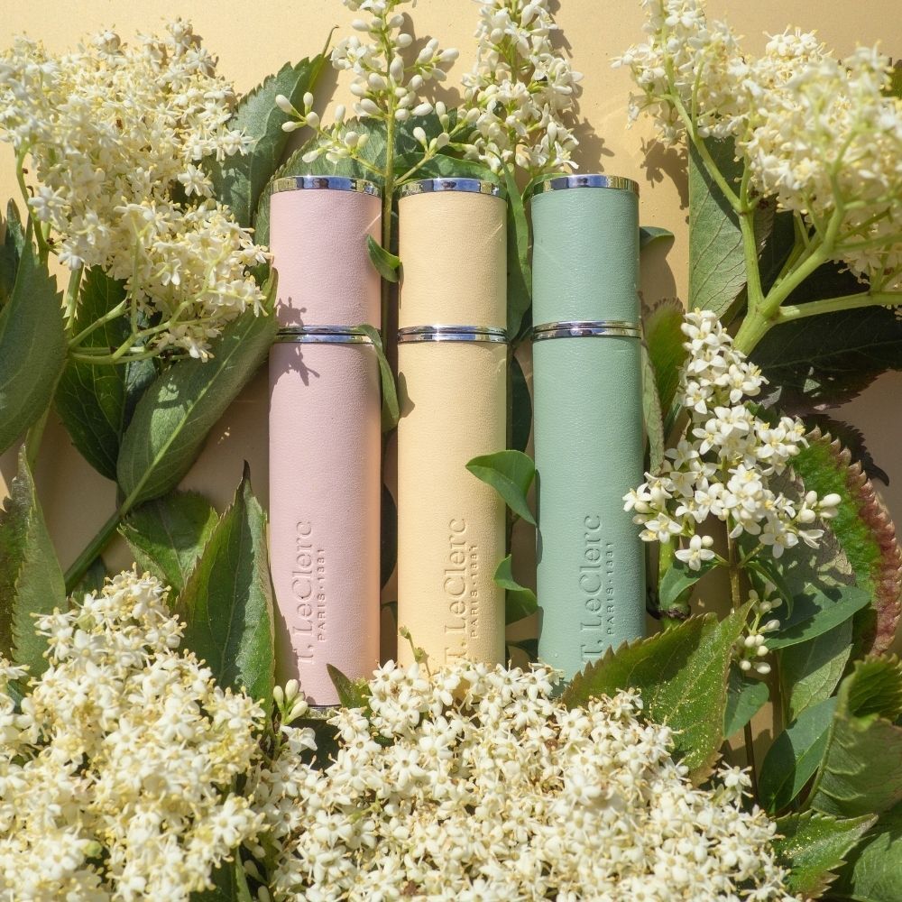 Drie T. LeClerc parfumverstuivers in roze, geel en groen, omringd door bloemen en bladeren.