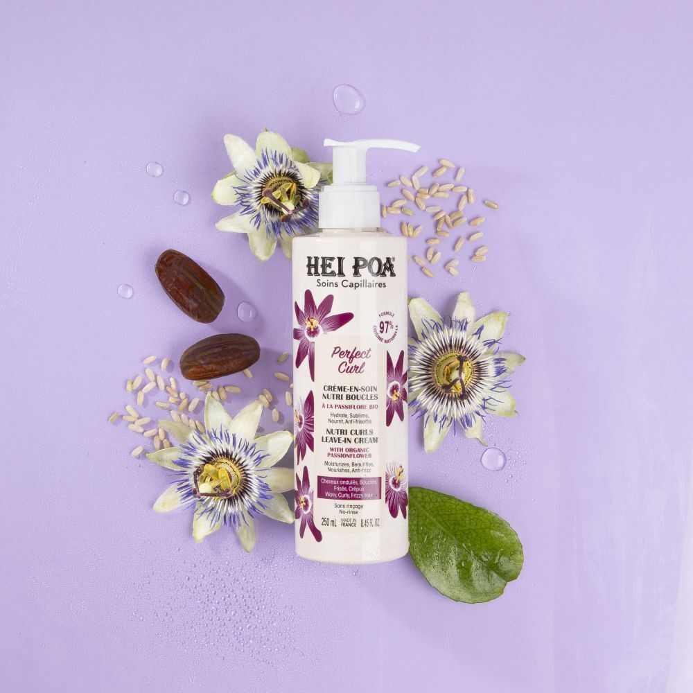 Een fles Hei Poa Perfect Curl Leave-in crème omringd door passiebloemen, bladeren en zaden op een paarse achtergrond.