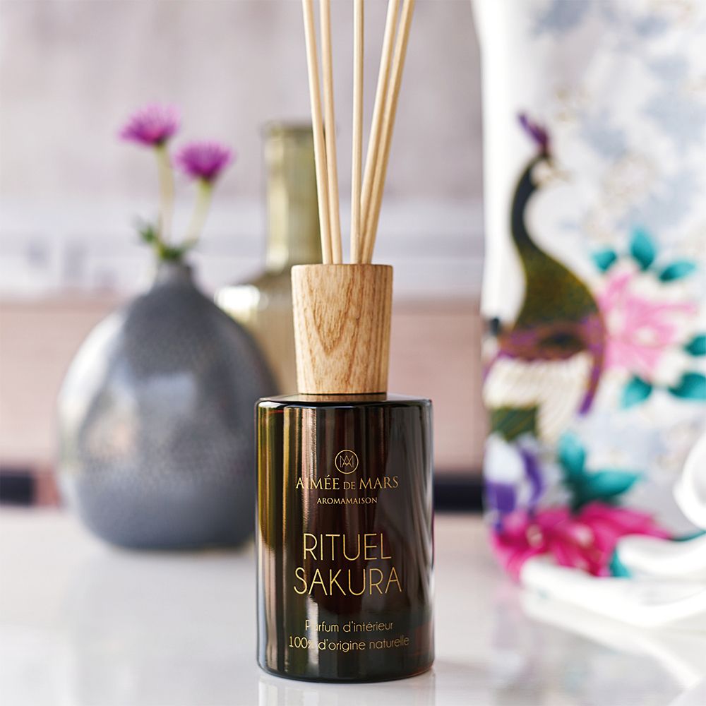 Bruine glazen fles met houten dop en stokjes. Opschrift: Rituel Sakura. Op de achtergrond vazen en bloemen.