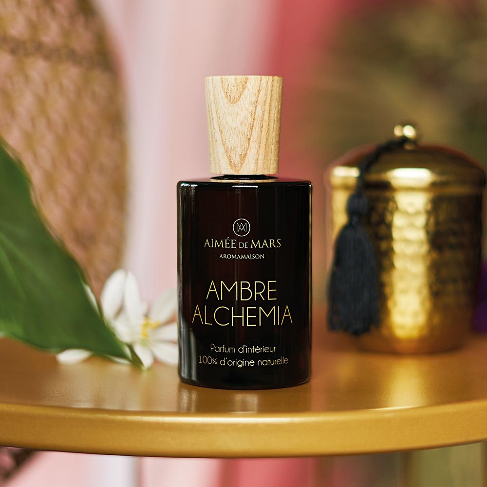 Bruine glazen fles met houten dop. Opschrift: AMBRE ALCHEMIA, Parfum d'intérieur, 100% d'origine naturelle. Op gouden tafel.