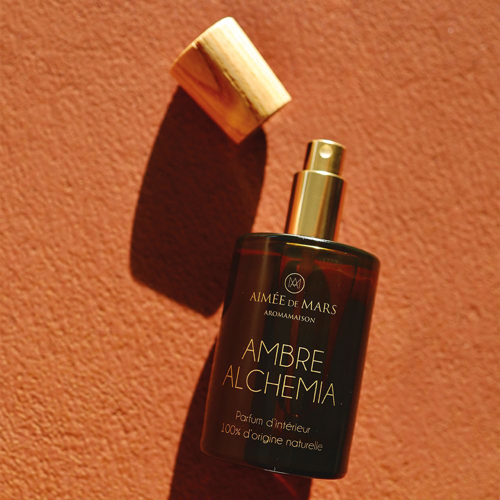 Bruine glazen fles met houten dop, deksel ernaast. Opschrift: AMBRE ALCHEMIA, Parfum d'intérieur, 100% d'origine naturelle.