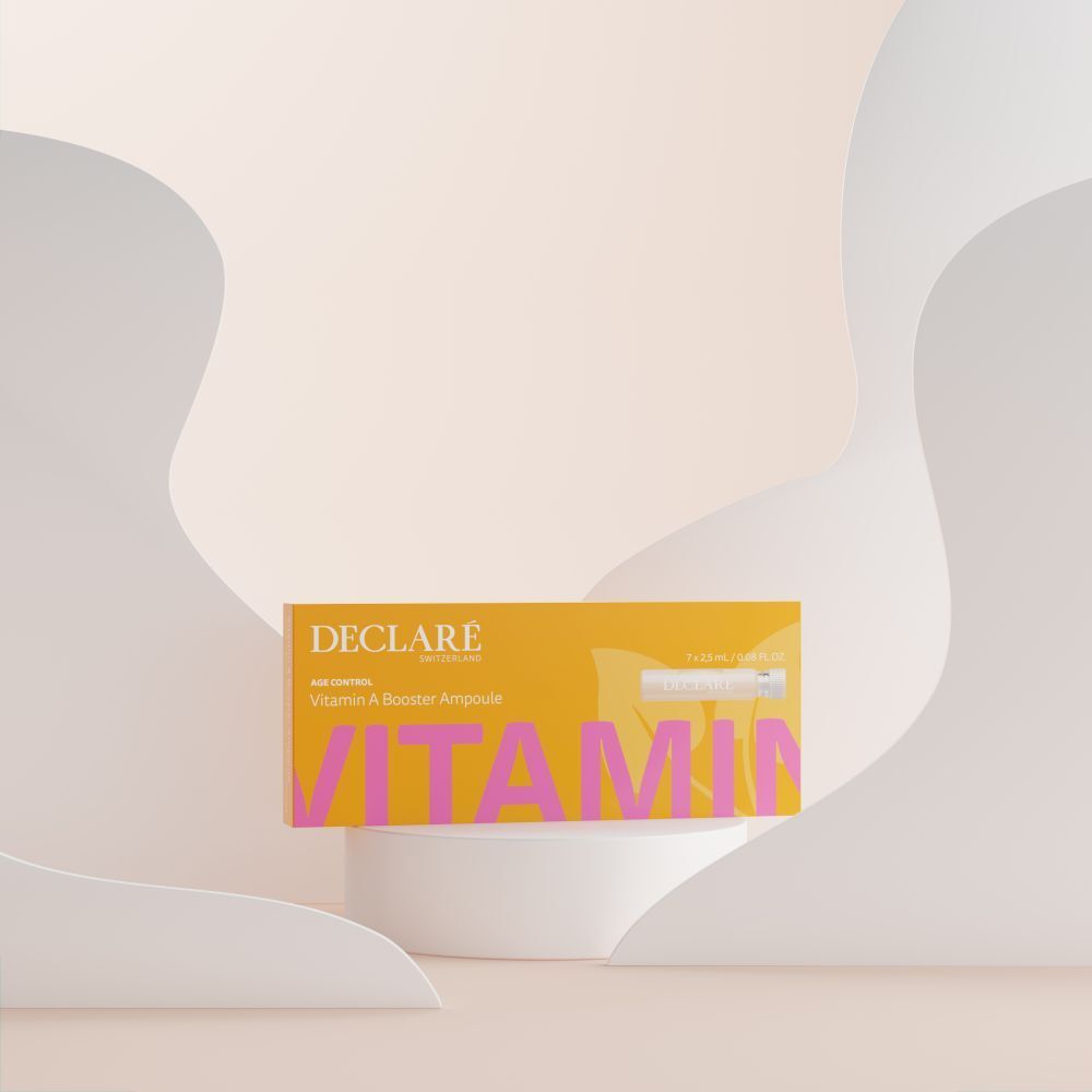 Gele doos DECLARÉ Vitamin A Booster Ampulle op sokkel. Roze tekst "VITAMIN". Achtergrond met vormen.