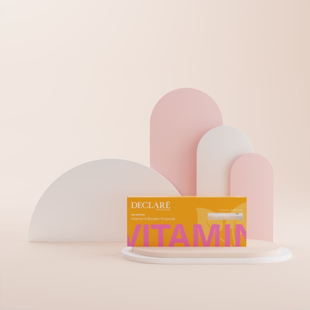 Gele doos DECLARÉ Vitamin A Booster Ampulle op sokkel. Roze tekst "VITAMIN". Achtergrond met vormen.
