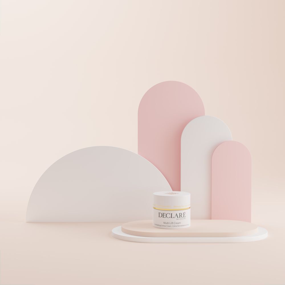 Pot de crème sur piédestal. DECLARÉ Multi Lift Cream. Fond avec formes blanches et roses.