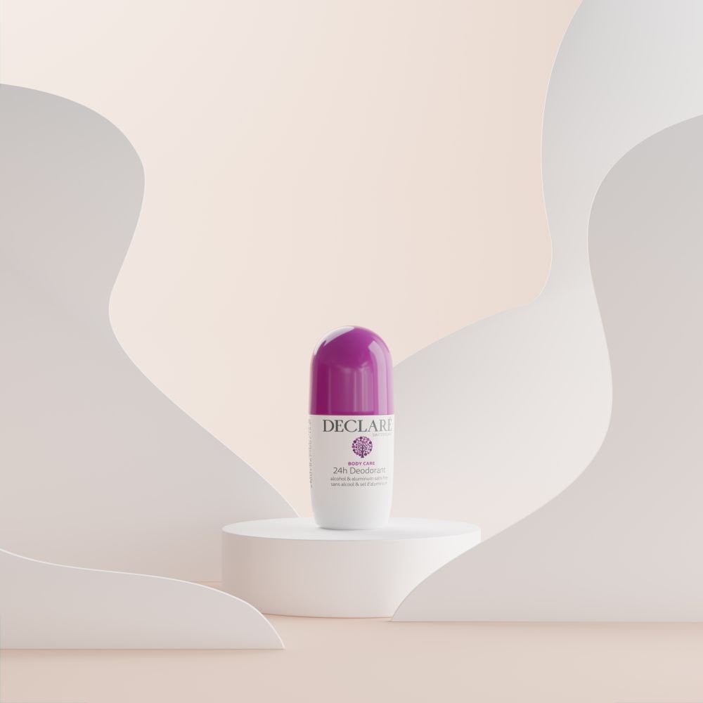 Déodorant roll-on sur socle. Boîtier blanc, bouchon violet. DECLARÉ, 24h Deodorant. Fond: formes blanches incurvées.