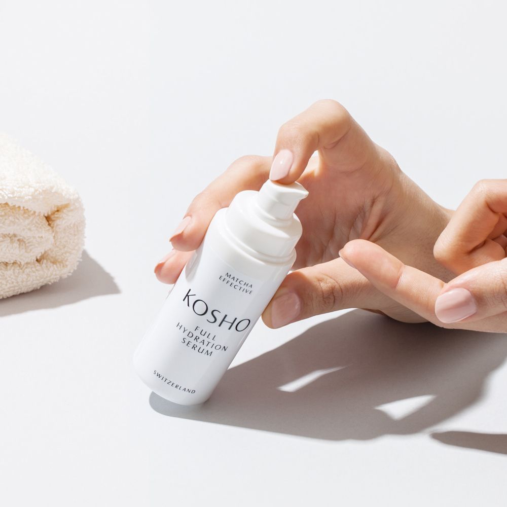 Witte serumfles met opschrift KOSHO Full Hydration Serum. Handen houden fles en handdoek vast.
