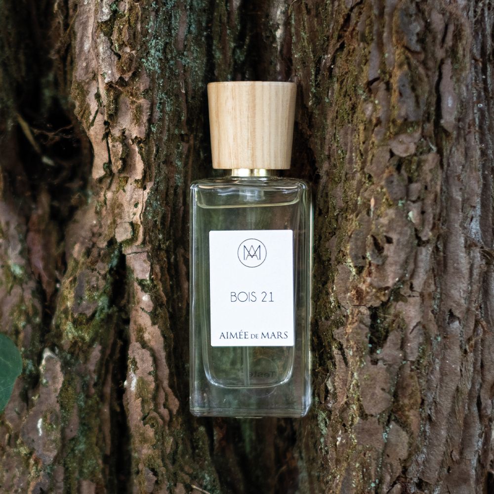 Flacon en verre avec bouchon en bois devant écorce d'arbre. Étiquette avec logo, texte « Bois 21 » et « Aimée de Mars ». Liquide visible.