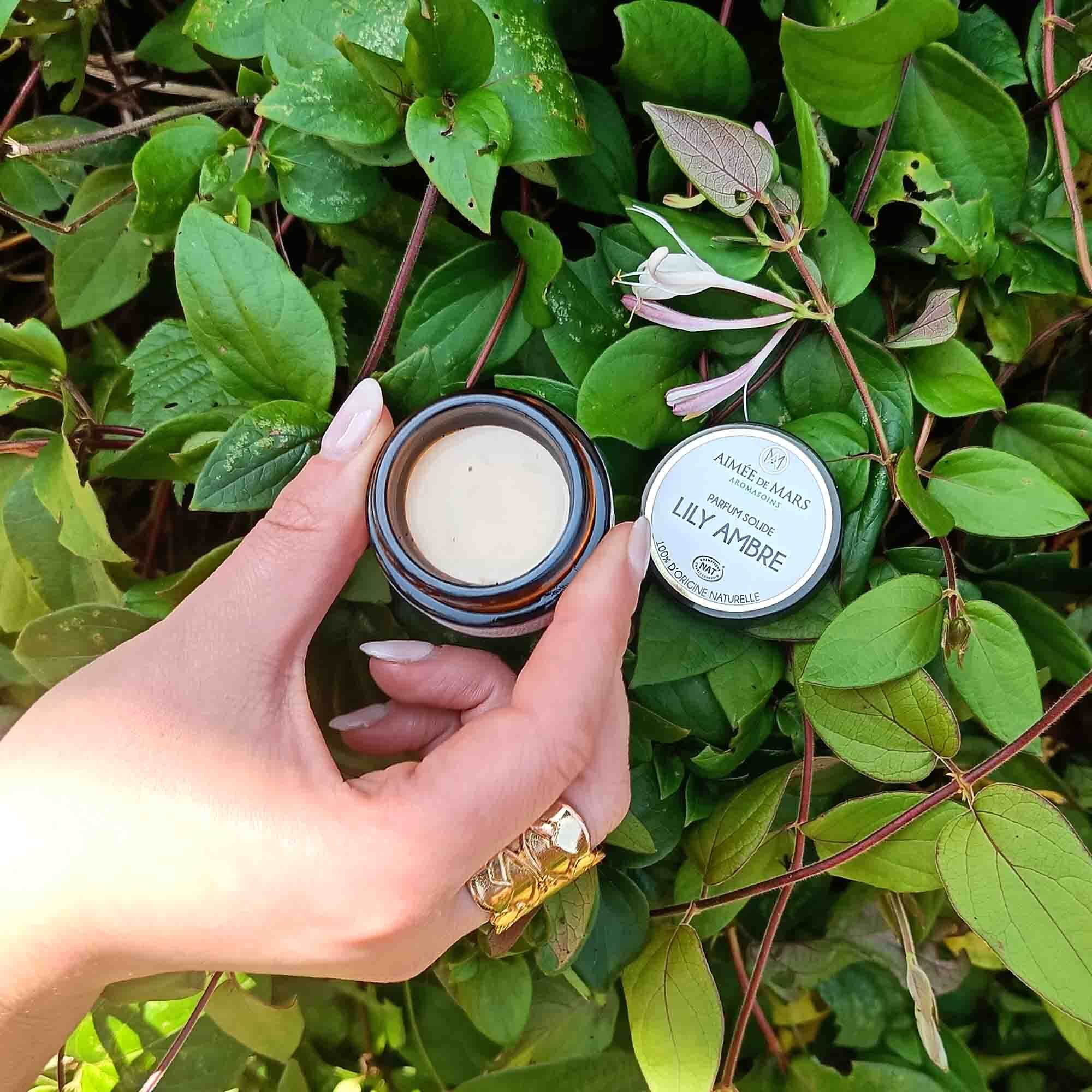 Hand houdt een geopend bruin potje met crème-kleurige inhoud en een deksel vast. Opschrift "LILY AMBRE".