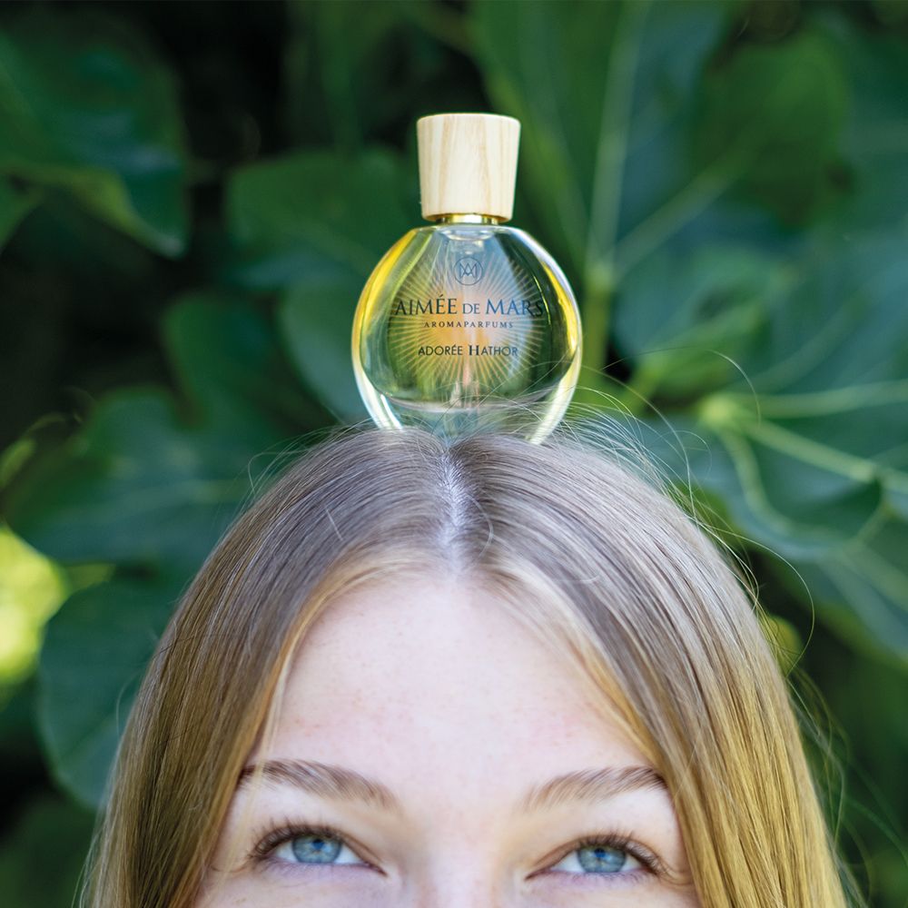 Femme avec flacon sur la tête. Flacon: rond, verre, bouchon bois. Inscription: AIMÉE DE MARS, ADORÉE HATHOR.