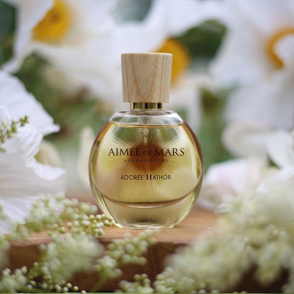 Flacon sur planche de bois, entouré de fleurs. Flacon: rond, verre, bouchon bois. Inscription: AIMÉE DE MARS, ADORÉE HATHOR.