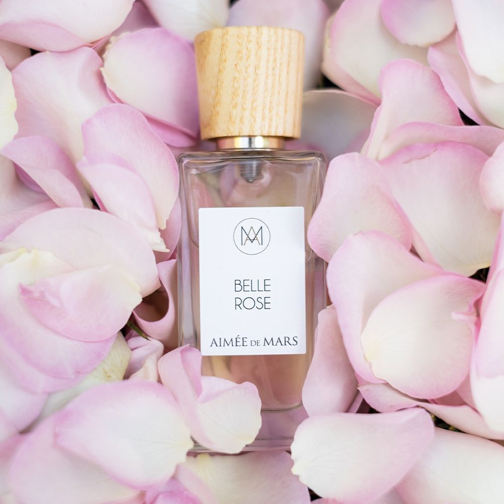Flacon Aimée de Mars Belle Rose Eau de Parfum entouré de pétales de rose roses. Bouteille rectangulaire en verre, bouchon en bois.