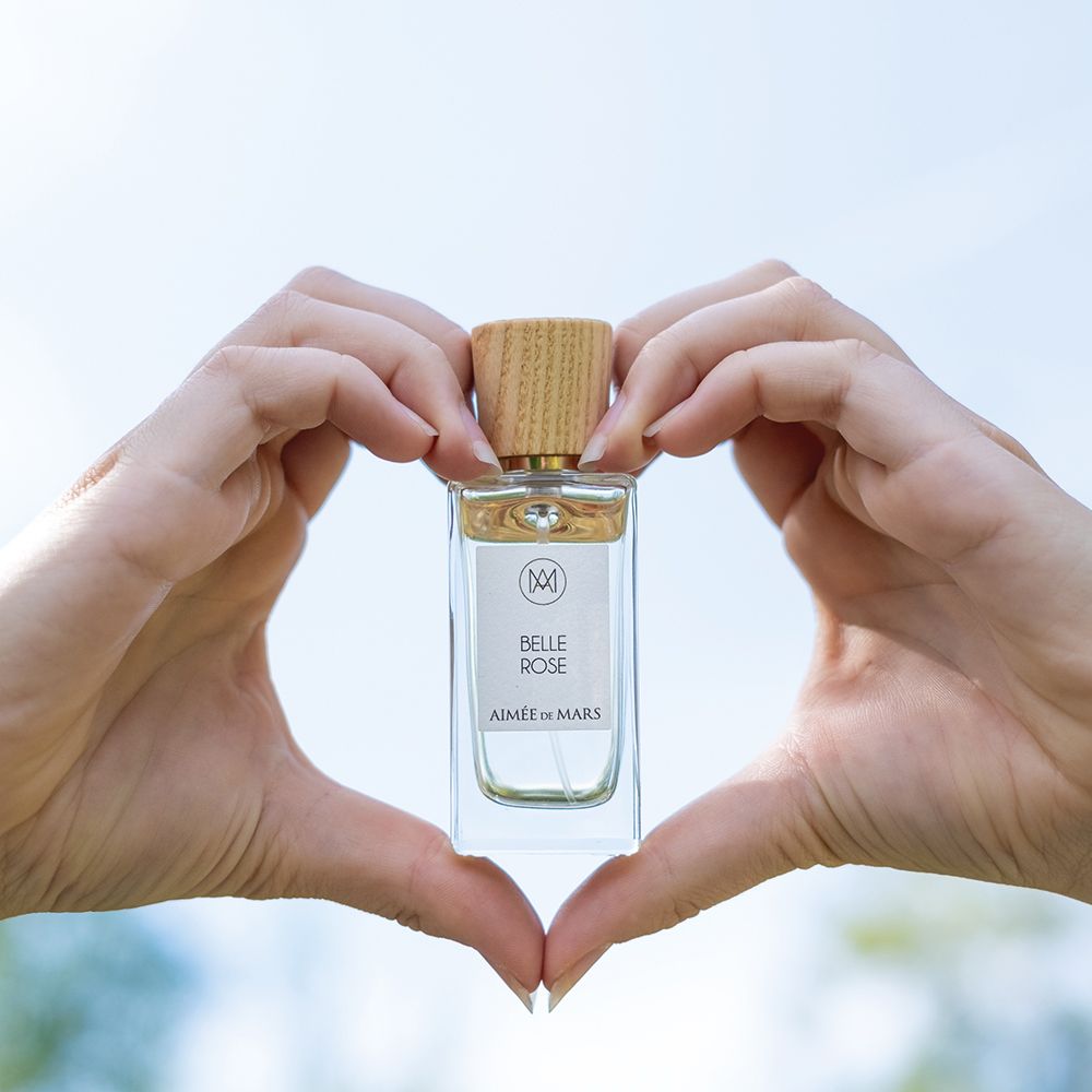 Flacon Aimée de Mars Belle Rose Eau de Parfum tenu par des mains en forme de cœur. Bouteille rectangulaire en verre, bouchon en bois.