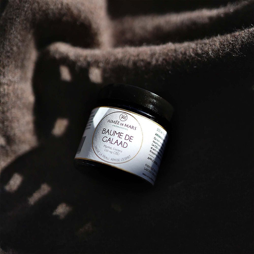 Pot met zwarte deksel op bruine achtergrond. Etiket: AIMÉE DE MARS, Baume de Galaad, Myrrhe, Encens, 100 mg CBD.