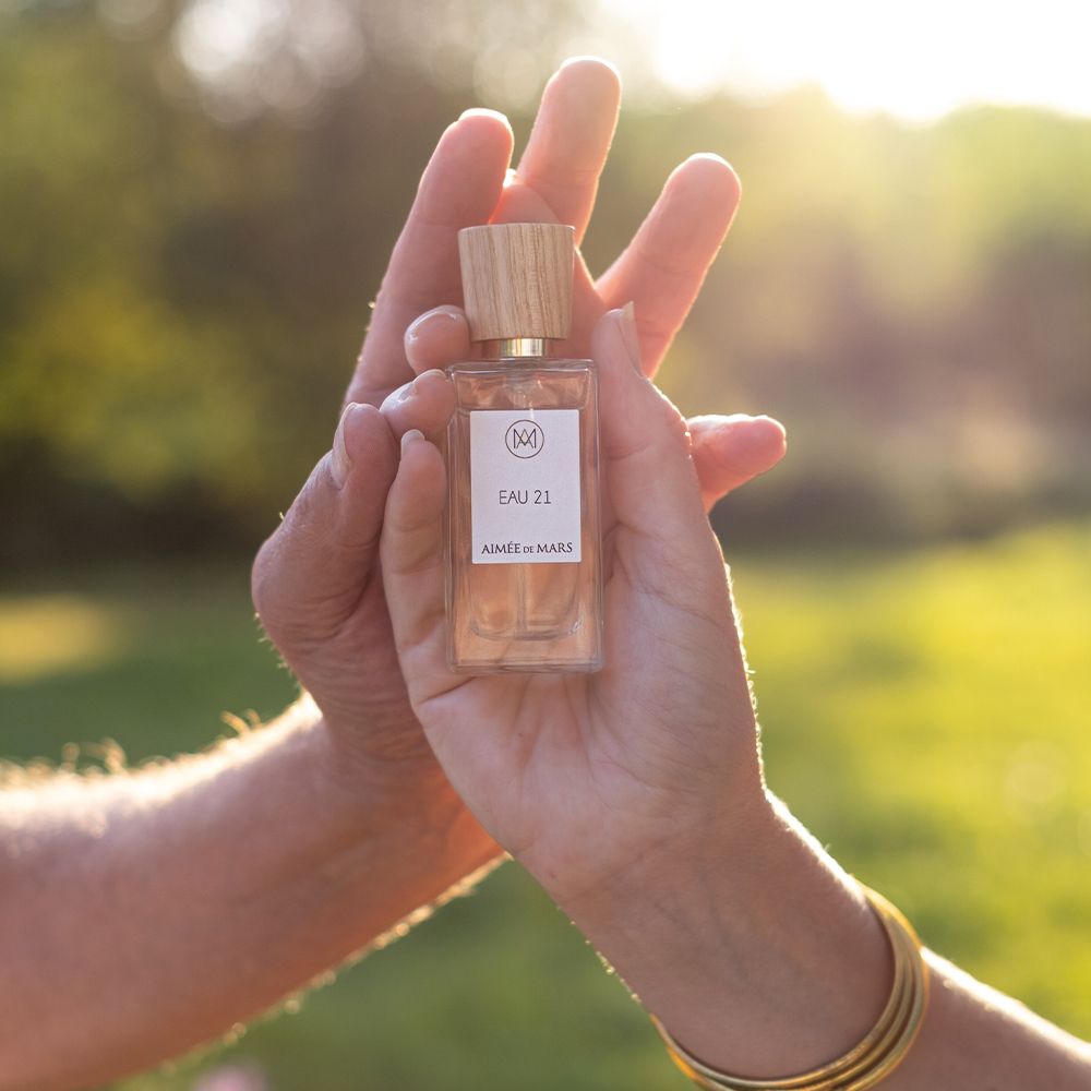 Parfumfles in handen. Houten dop, label met logo, "EAU 21" en "Aimée de Mars".