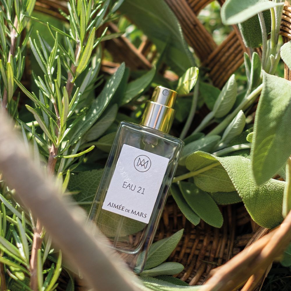 Parfumfles omringd door kruiden. Label met logo, "EAU 21" en "Aimée de Mars".