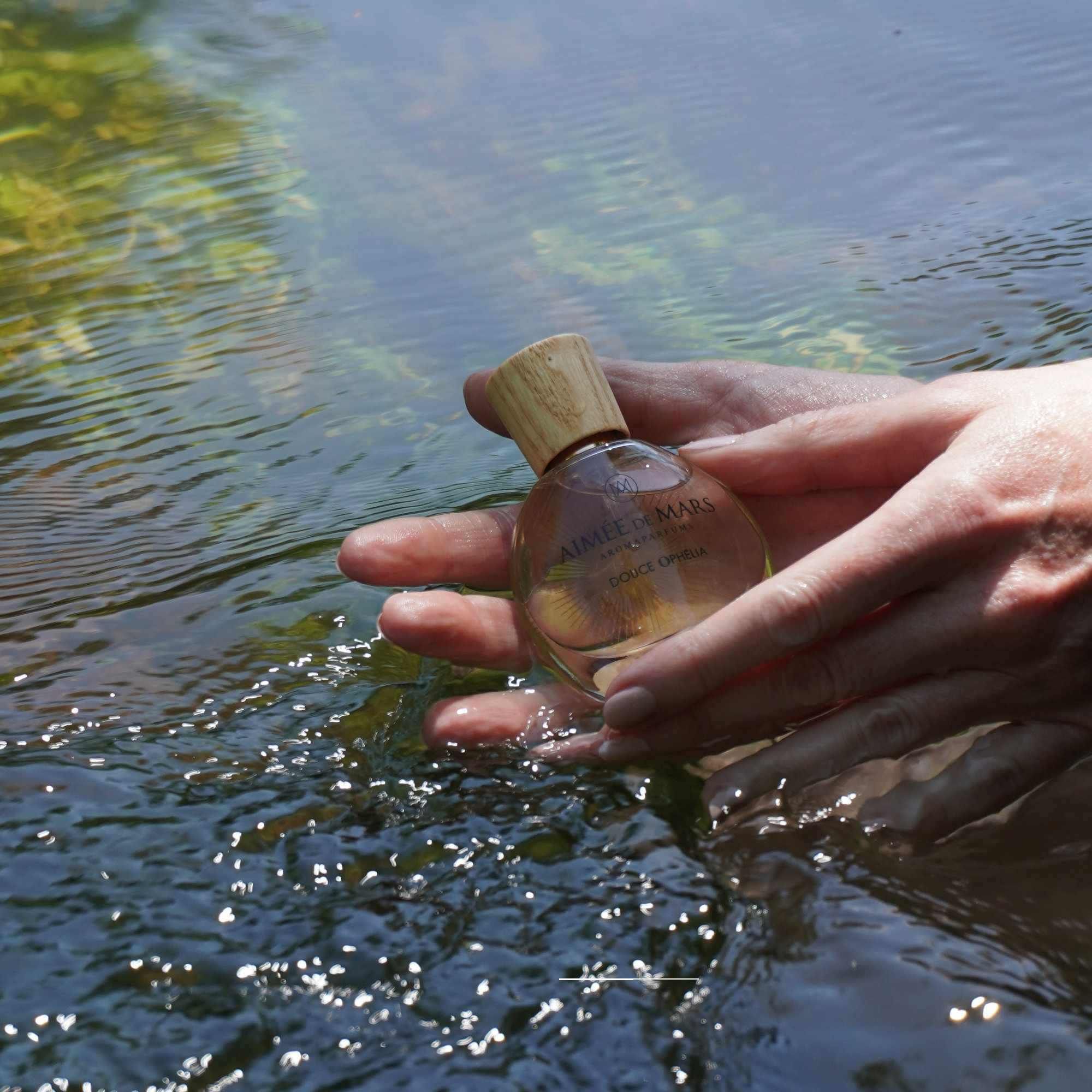 Mains tenant un flacon en verre dans l'eau. Bouchon en bois visible. Inscription : Aimée de Mars, Douce Ophélia.