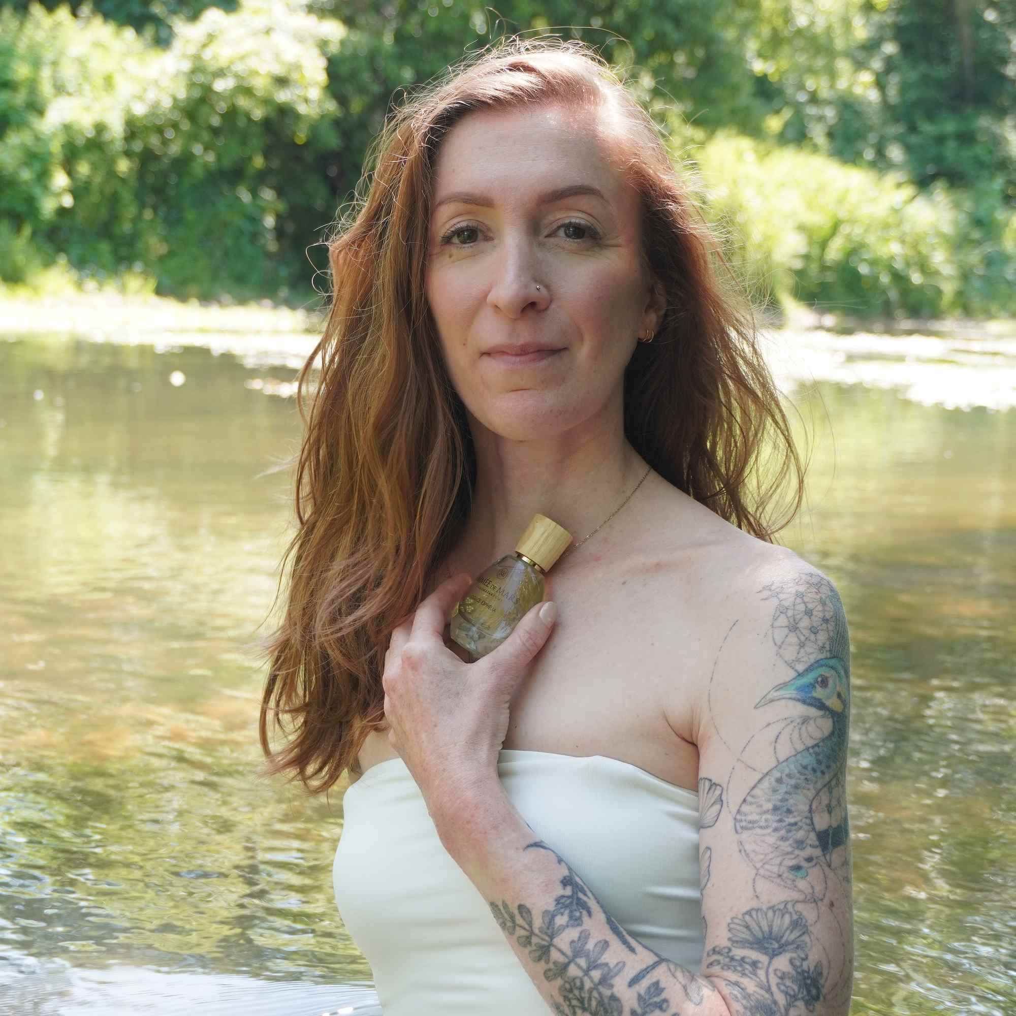 Femme tenant un flacon en verre. Bouchon en bois visible. Inscription : Aimée de Mars, Douce Ophélia. Tatouage sur le bras.