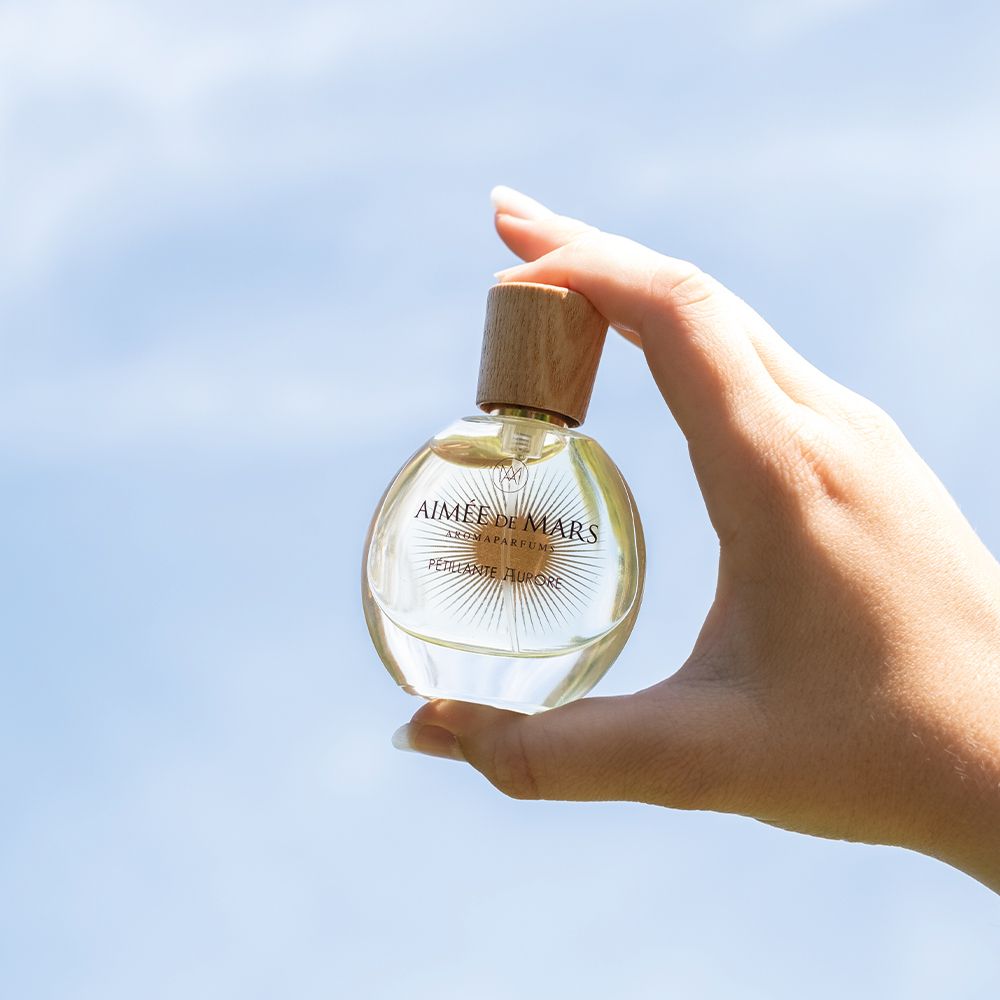 Main tenant un flacon de parfum rond. Inscription : Aimée de Mars, Pétillante Aurore. Ciel bleu en arrière-plan.