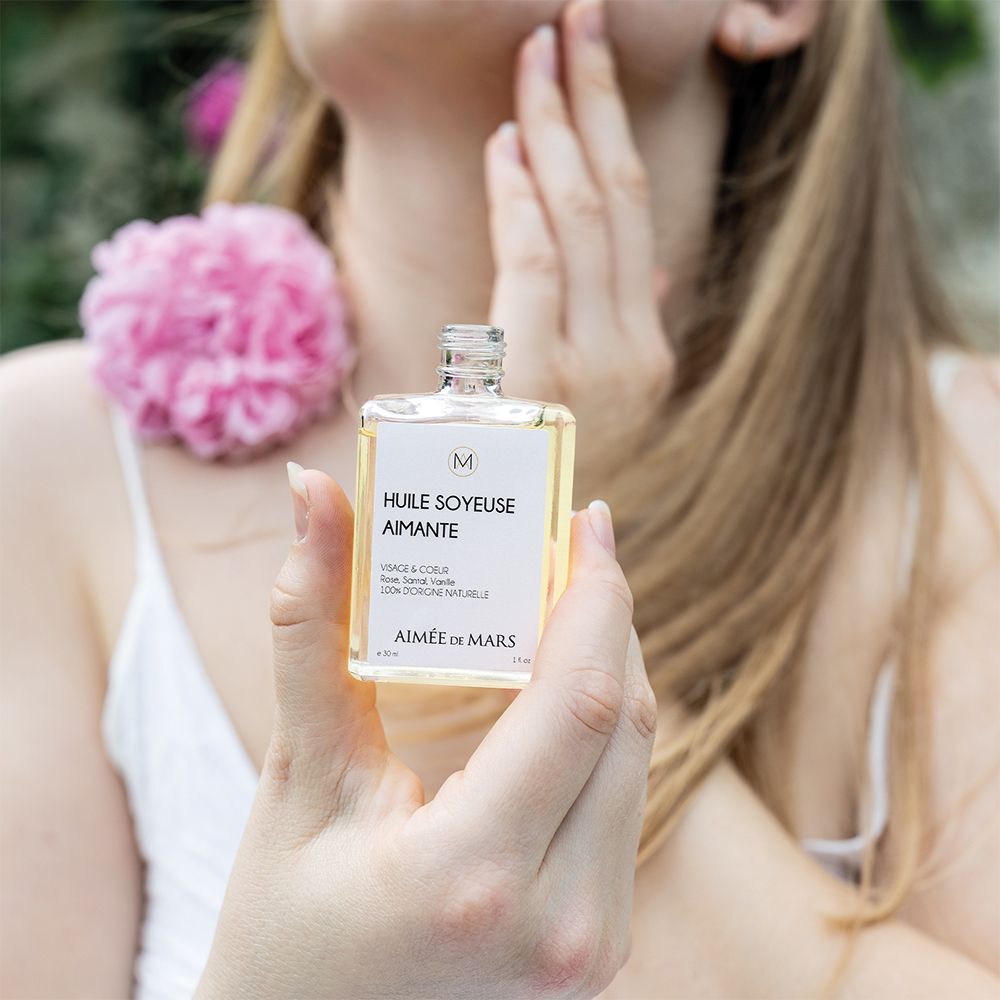 Vrouw houdt een glazen fles vast. Opschrift: Huile Soyeuse Aimante, Aimée de Mars. Roze bloem op de achtergrond.