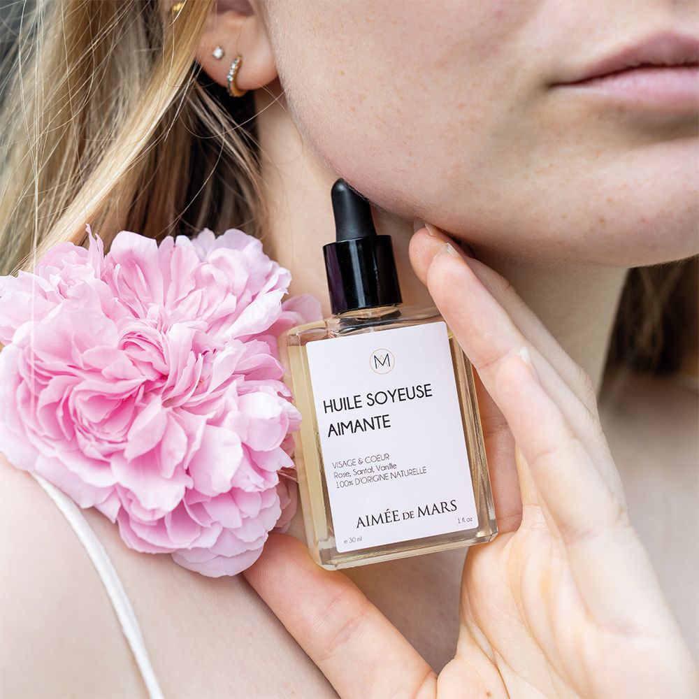 Glazen fles met zwarte druppelaar. Opschrift: Huile Soyeuse Aimante, Aimée de Mars. Roze bloem naast de vrouw.