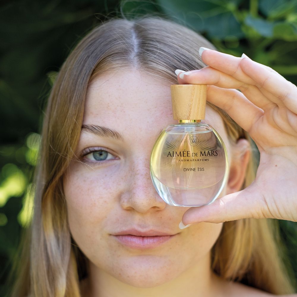 Vrouw houdt fles voor gezicht. Fles: rond, glas, houten dop. Opschrift: Aimée de Mars, Divine Isis. Goudkleurige vloeistof.