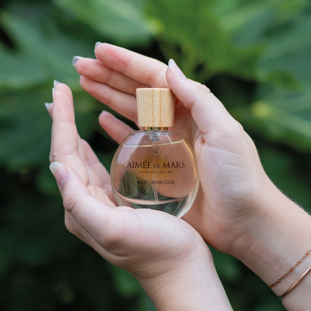 Flacon de parfum tenu dans les mains. Inscription : Aimée de Mars, Belle Aphrodite. Bouchon en bois. Liquide doré.