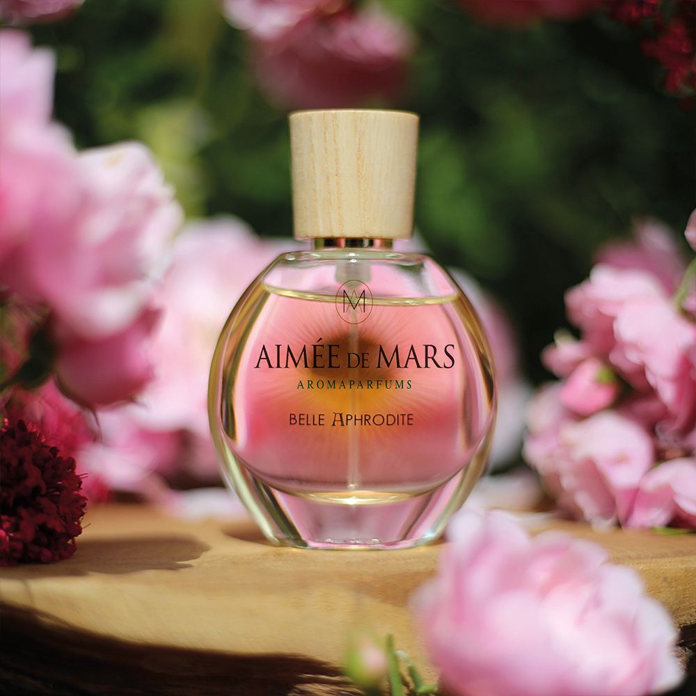 Flacon de parfum devant des roses. Inscription : Aimée de Mars, Belle Aphrodite. Bouchon en bois. Liquide doré.