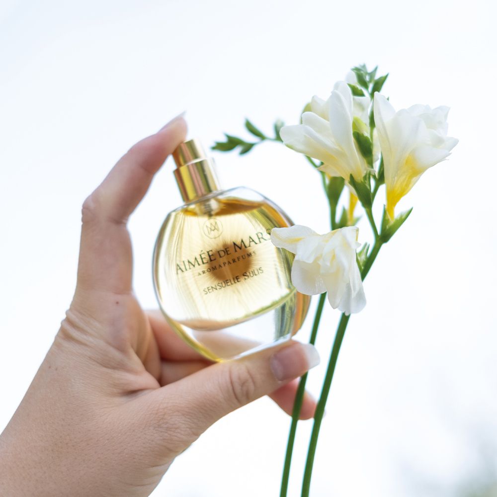 Main tenant un flacon rond en verre avec liquide doré. Inscription : Aimée de Mars Aromaparfums, Sensuelle Sulis.