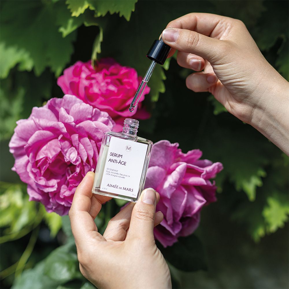 Handen houden flesje en pipet boven rozen. Flesje met "SERUM ANTI-ÂGE" en "AIMÉE DE MARS".