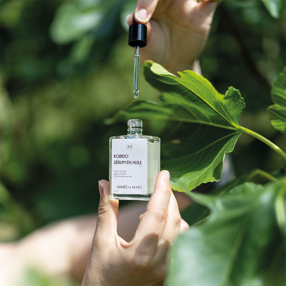 Handen houden een glazen fles met olie-serum en druppelaar boven groene bladeren. Druppels vallen.