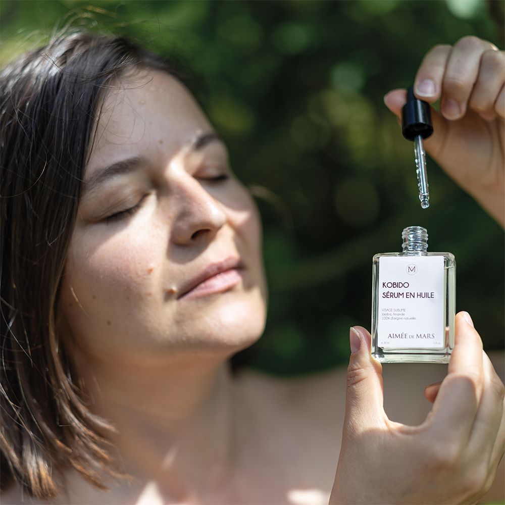 Vrouw houdt een glazen fles met olie-serum en druppelaar vast. Ontspannen gezicht, ogen dicht. Groene achtergrond.