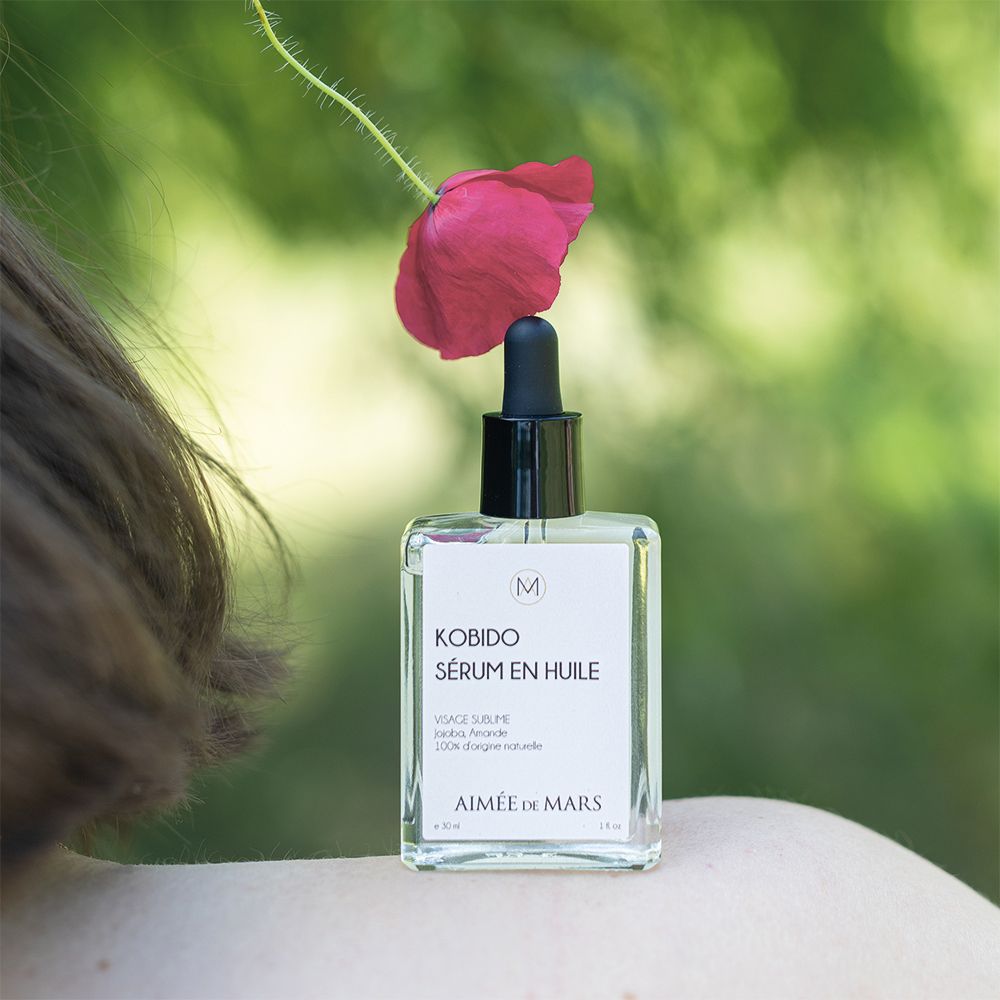 Glazen fles met olie-serum en druppelaar, geplaatst naast een rode bloem. Schouder van een persoon op de achtergrond.