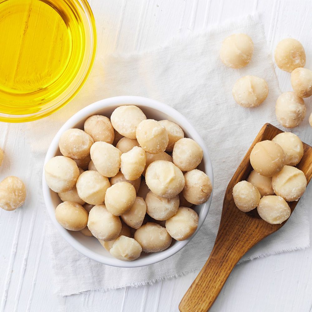 Bol blanc avec noix de macadamia, cuillère en bois avec noix, à côté huile dans un bol en verre.