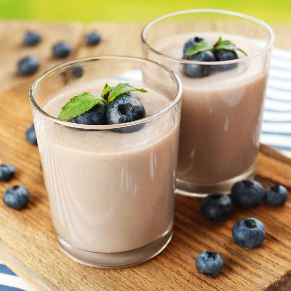 Twee glazen met paarse shake, bosbessen en munt op een houten plank. Bosbessen verspreid.