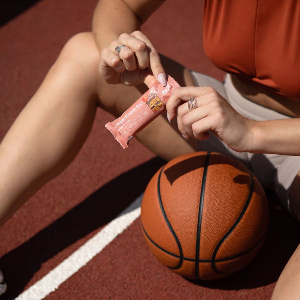 Persoon houdt een eiwitreep vast. Opschrift: Fit Vegan Protein Bar. Basketbal en sportveld op de achtergrond.