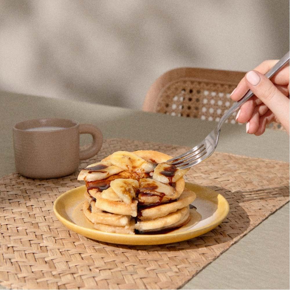 Pannenkoeken op een bord met bananenschijfjes en chocoladesaus. Een hand houdt een vork vast.