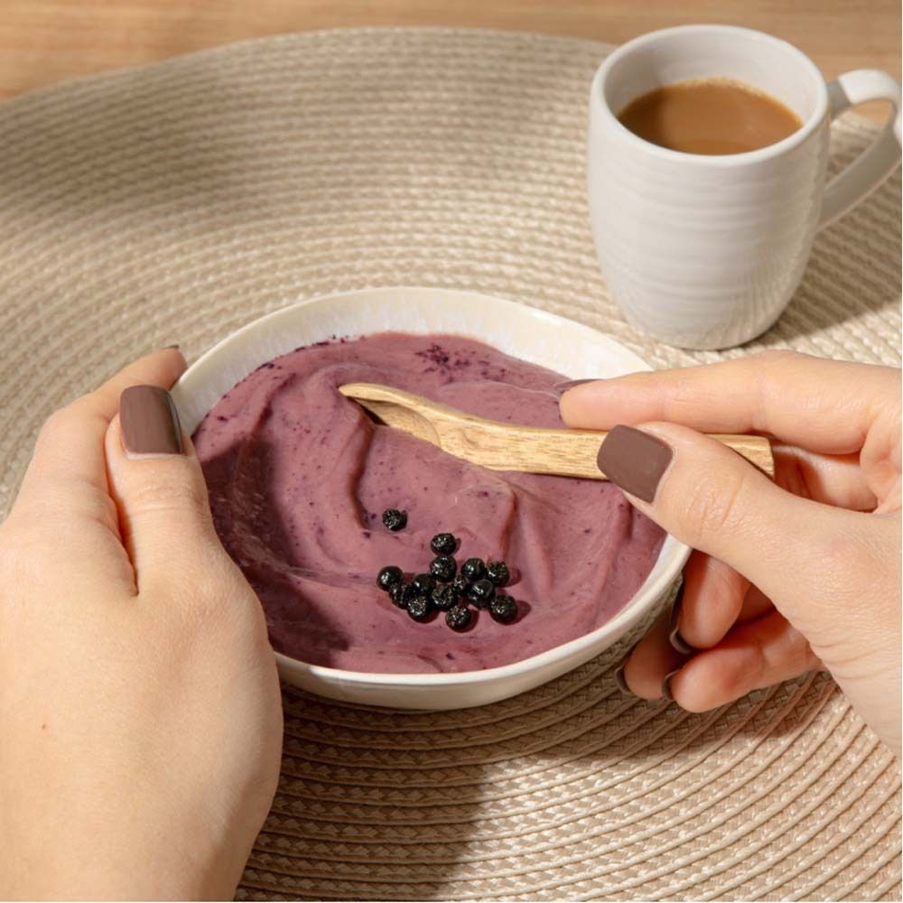 Gros plan sur un bol de smoothie violet, baies d'aronia et une cuillère en bois. Une tasse de café est à côté. Mains tenant le bol.
