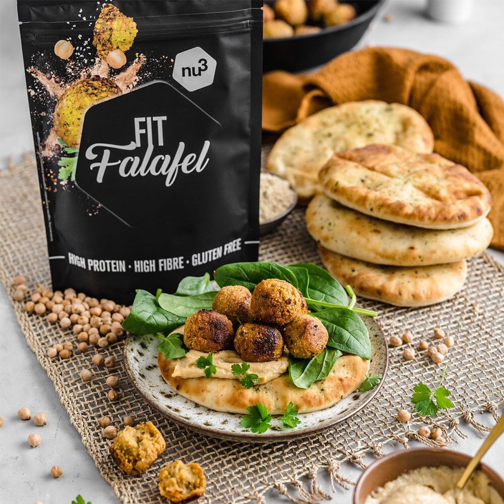 nu3 Fit Falafel-verpakking naast een bord met falafel, hummus en brood. Kikkererwten en stukjes brood liggen ernaast.