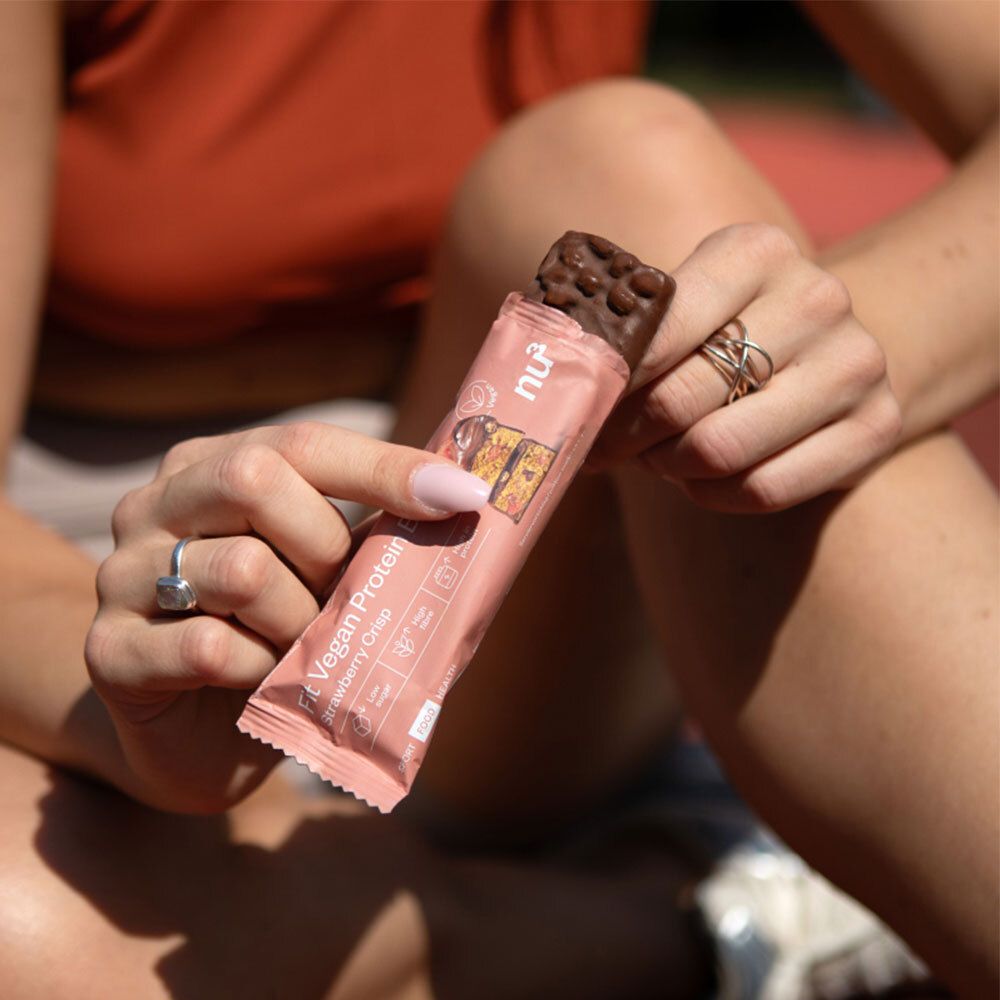 Personne ouvrant une barre protéinée dans un emballage rose. Barre avec enrobage chocolat visible. Nom et logo du produit.