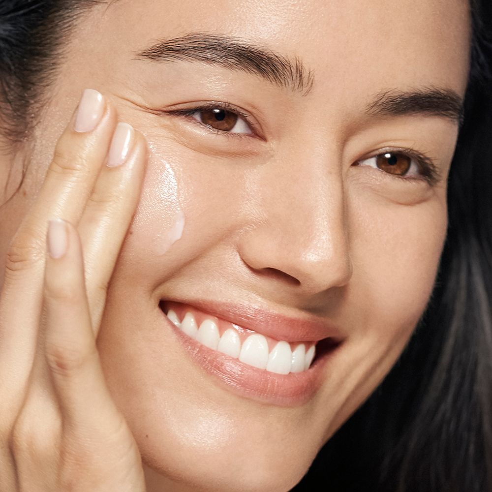 Gros plan d'une femme appliquant de la crème sur son visage. Elle sourit.