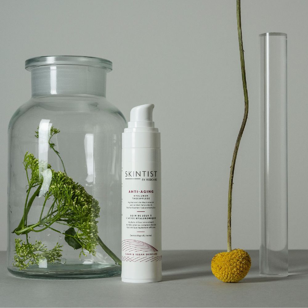 Un flacon blanc SKINTIST ANTI-AGING Hyaluron Tagespflege à côté d'une décoration. Texte en allemand et en anglais.