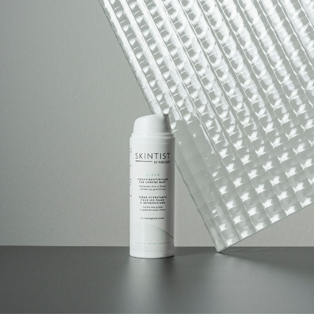 Witte fles SKINTIST CLEAR Feuchtigkeitspflege. Achtergrond: grijs oppervlak en transparant materiaal.