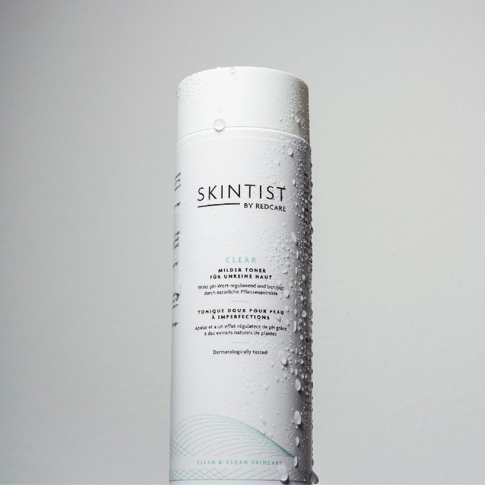 Een witte fles SKINTIST CLEAR Milder Toner met waterdruppels. Opschrift: Voor onzuivere huid. Dermatologisch getest.
