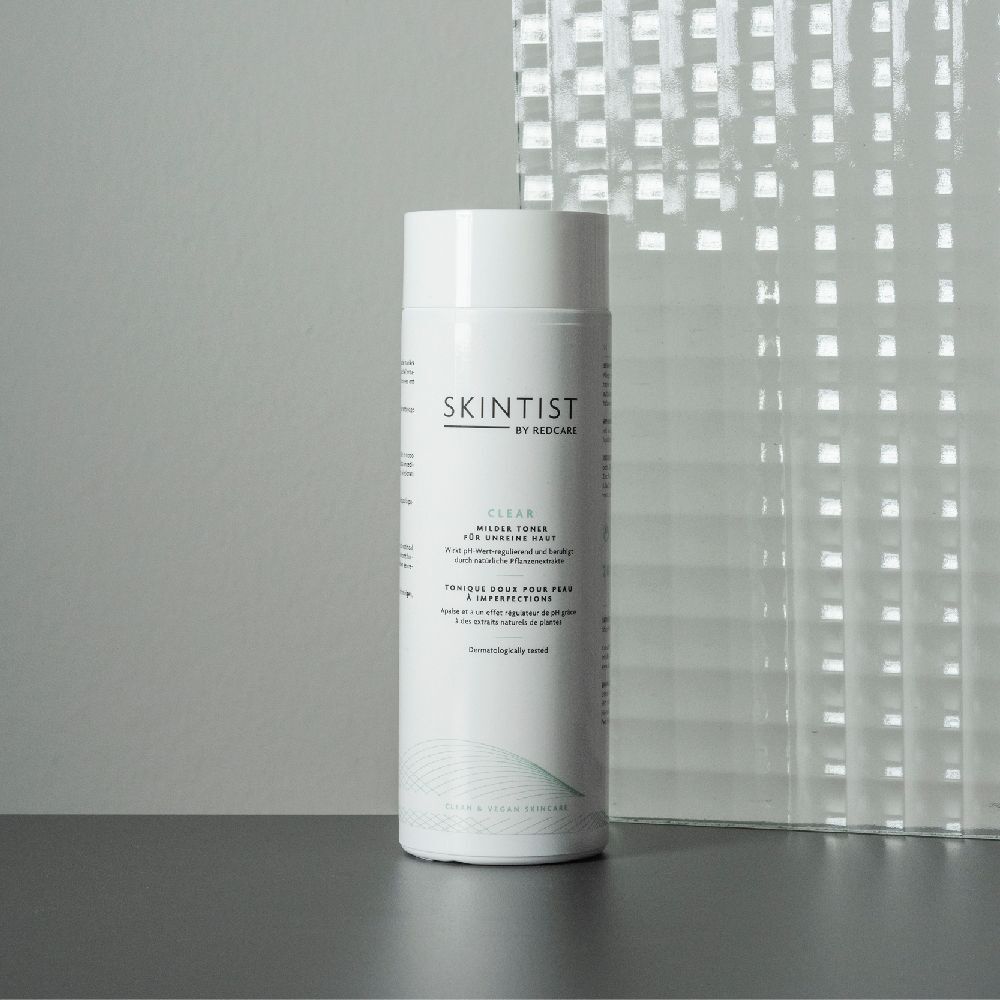 Een witte fles SKINTIST CLEAR Milder Toner op een tafel. Een glazen wand op de achtergrond. Opschrift: Voor onzuivere huid.