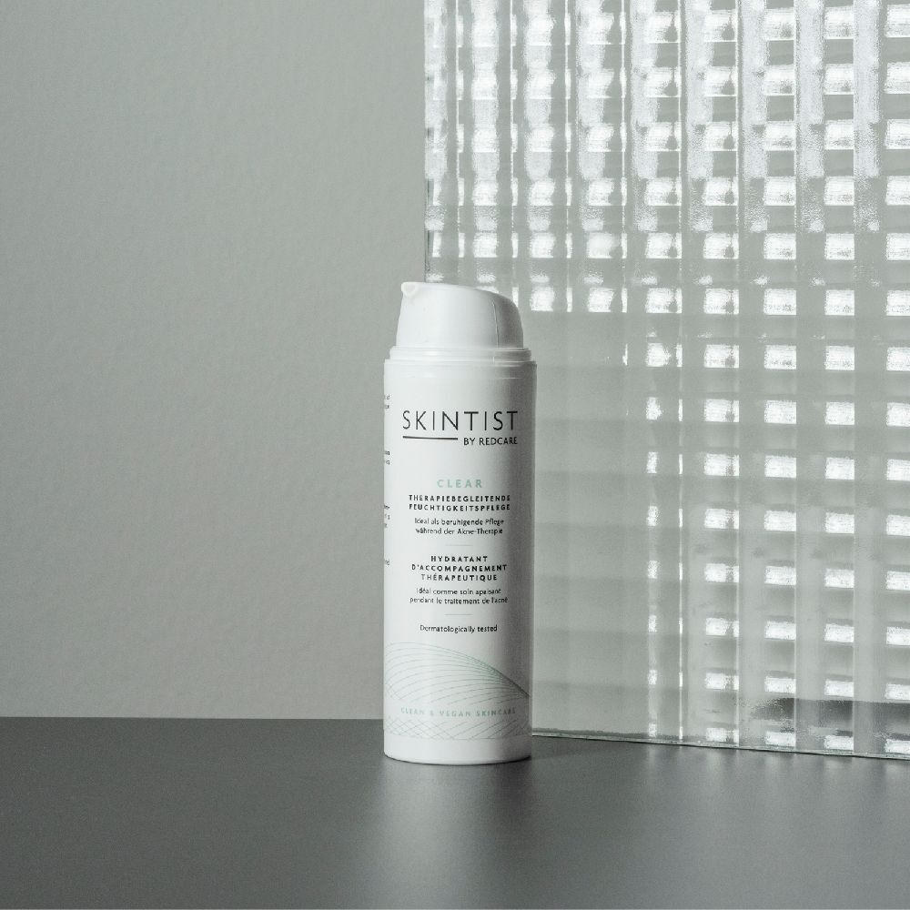 Witte fles SKINTIST CLEAR. Therapiebegeleidende Moisturizer. Staat voor een glazen wand.